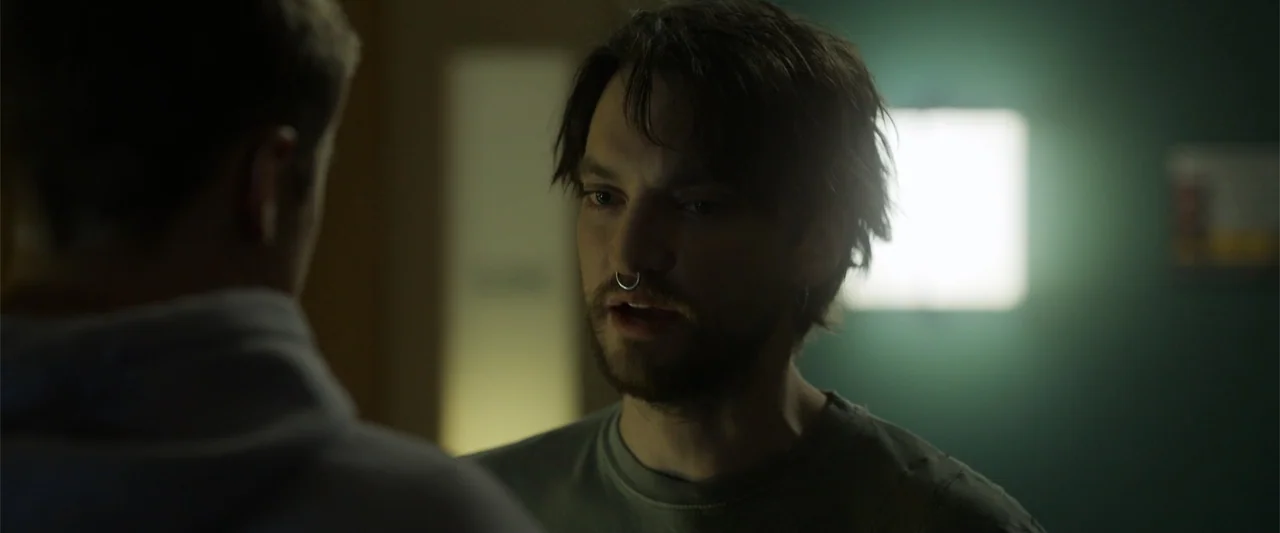 Final Destination Bloodlines (2025) movie still: medium — A dimly lit interior room with a glowing square light fixture on the back wall. A man with…