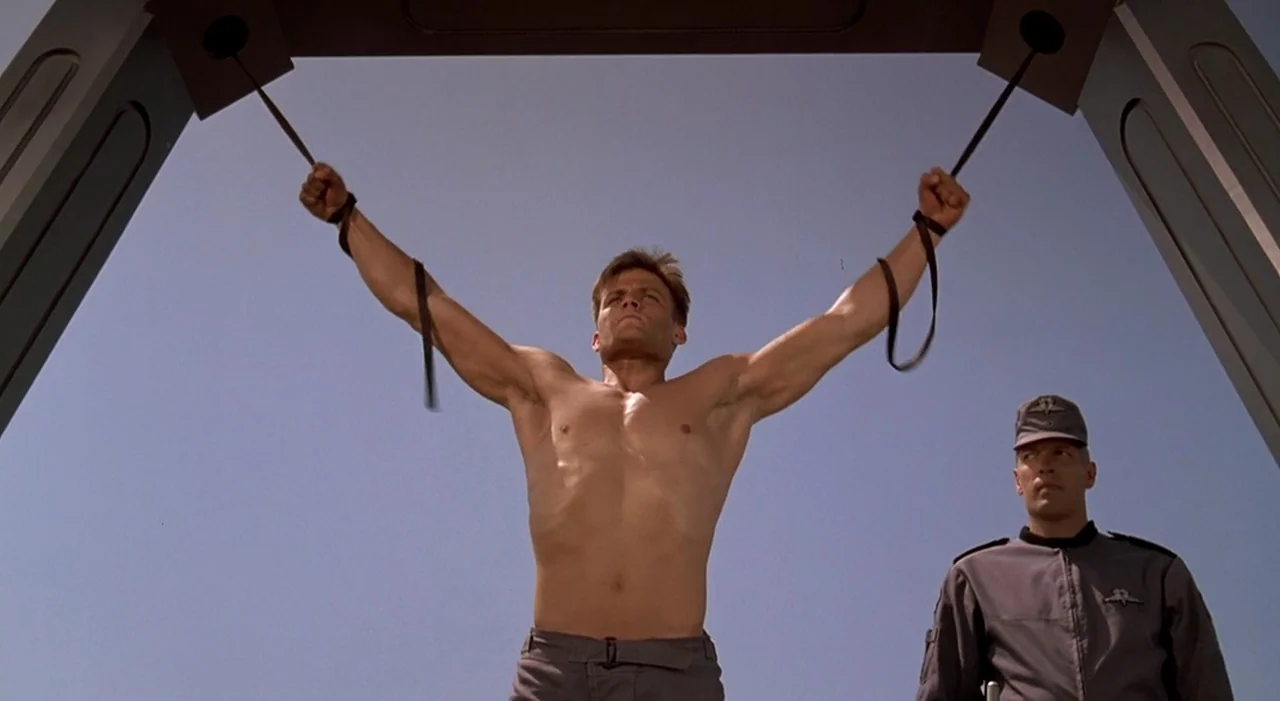Starship Troopers (1997) movie still: fullbody — A sunny outdoor training area features metal suspension structures against a clear sky. A man holds…