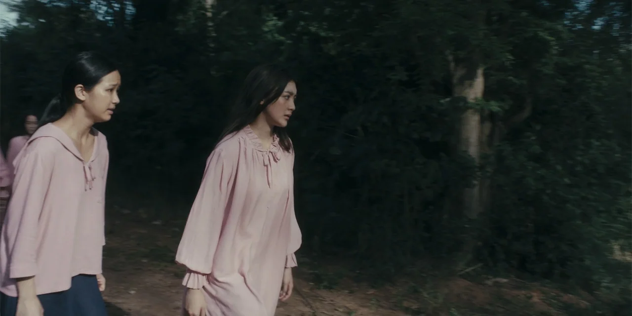 Host (2020) movie still: medium — A dense grove of trees with dark foliage provides a backdrop for the scene. Two women in matching pink clothing…