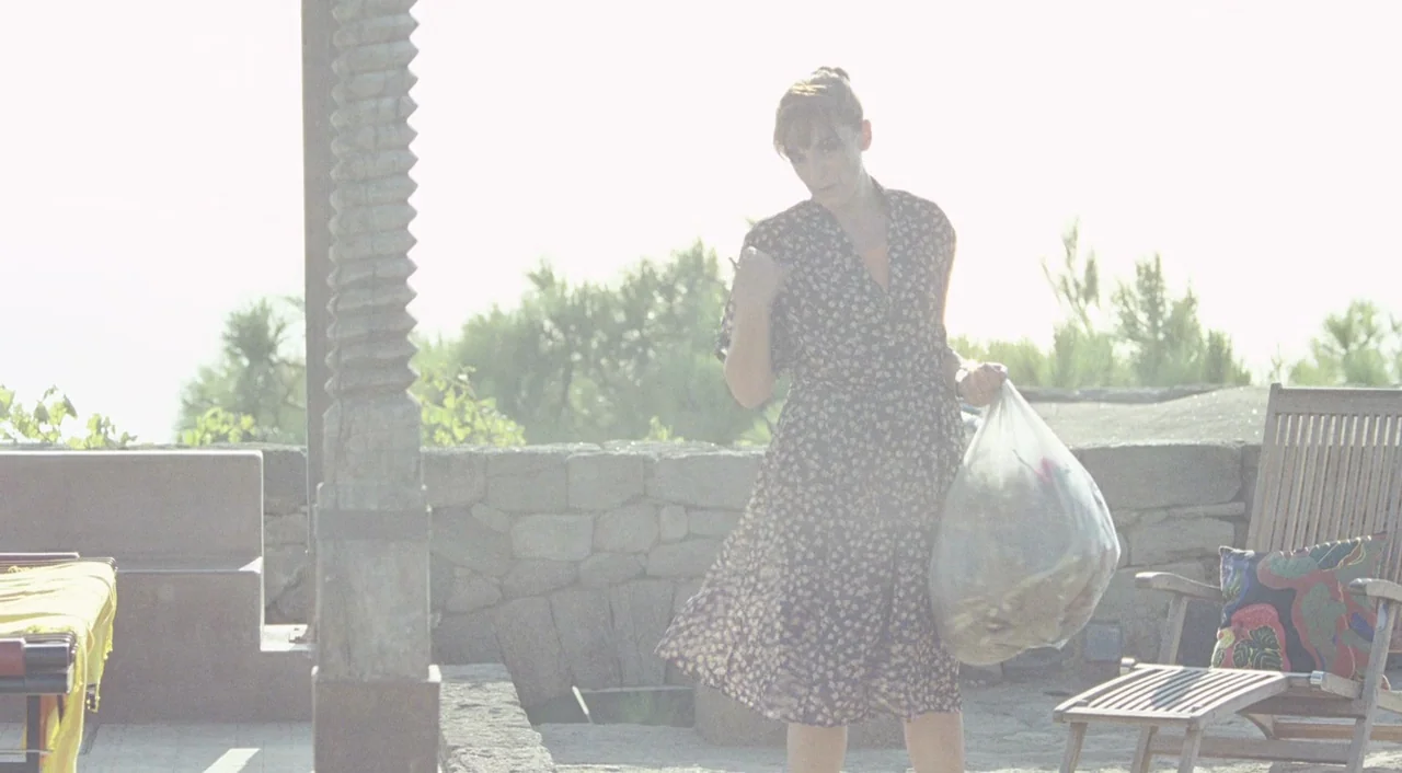 A Bigger Splash (2015) movie still: fullbody — A sun-drenched outdoor patio features a stone wall and wooden lounge furniture. A woman in a floral…