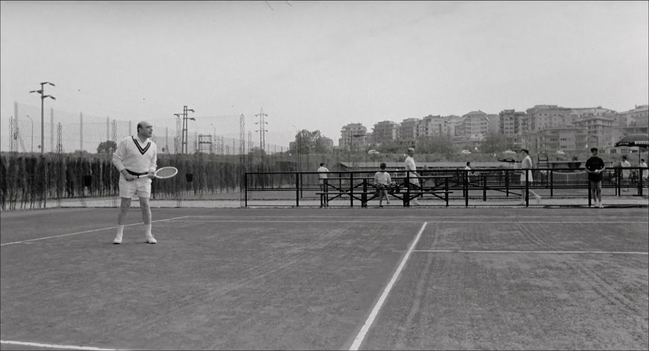Il Boom (1963) movie still: wide — An outdoor clay tennis court located near residential buildings under an overcast sky. A man in athletic wear…