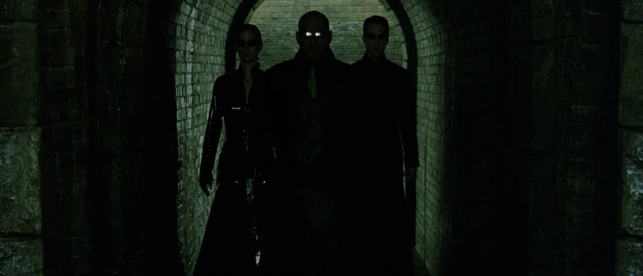 The Matrix Reloaded (2003) movie still: fullbody — A dark brick arched tunnel provides a narrow path for three figures walking forward. The group…