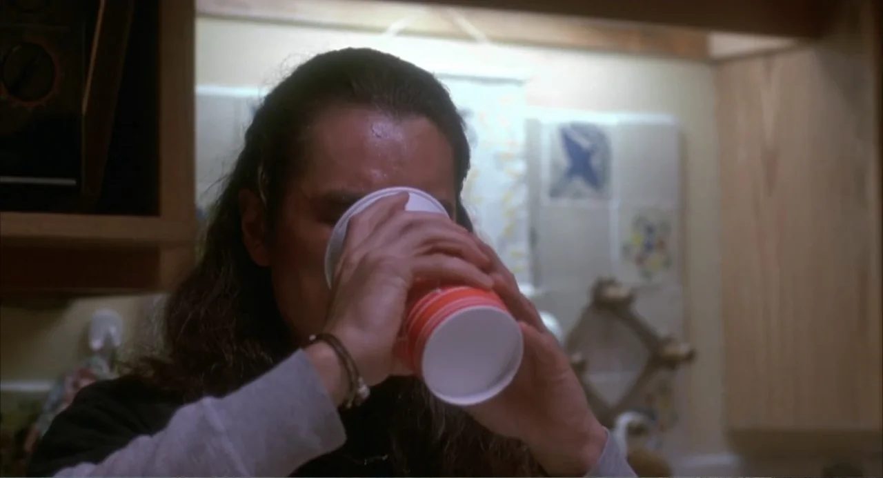3 Ninjas (1992) movie still: closeup — A dimly lit indoor kitchen space with wooden cabinets in the background. A person with long hair drinks from…