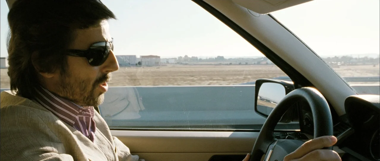 Rivals (1972) movie still: medium — A modern car cabin with beige trim and a black steering wheel. A man wearing sunglasses drives the vehicle…