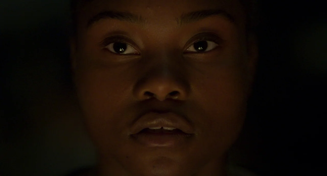 Zombi Child (2019) movie still: closeup — A close-up of a person's face against an entirely black, featureless background. The subject gazes…
