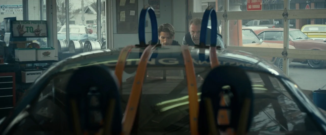 God Bless the Broken Road (2018) movie still: medium — An auto repair shop interior features a race car positioned centrally in the foreground. Two…