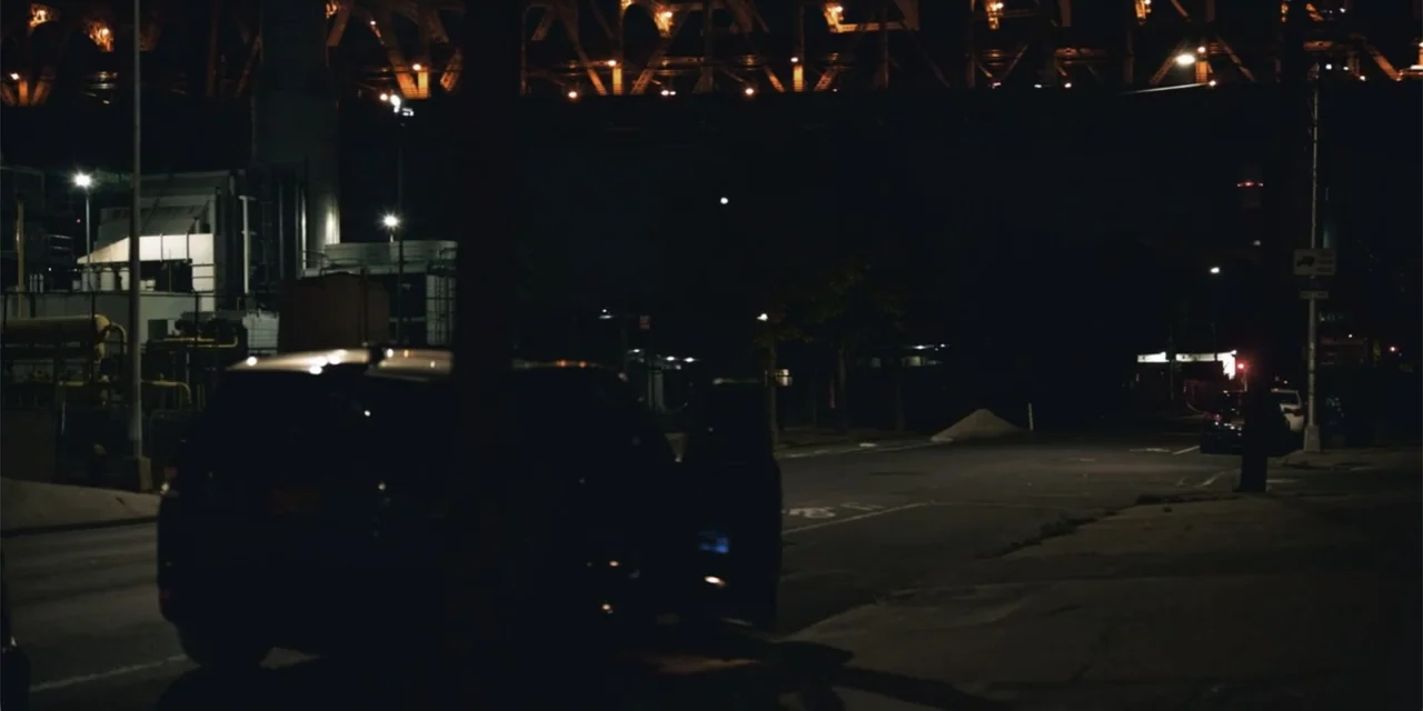 Equal Standard (2021) movie still: wide — An urban street at night passes under a large metal bridge structure. A dark vehicle is parked with its…