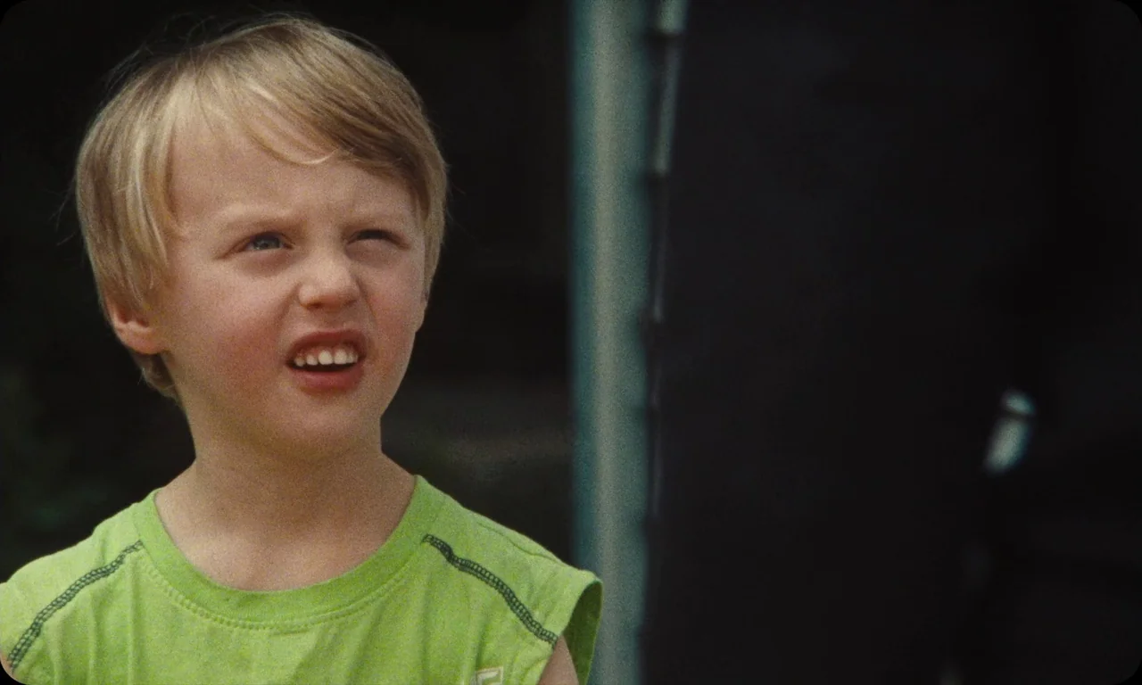 Vulcanizadora (2024) movie still: closeup — A close-up of a young boy standing in front of a metal chain-link fence. The boy squinting as he looks…