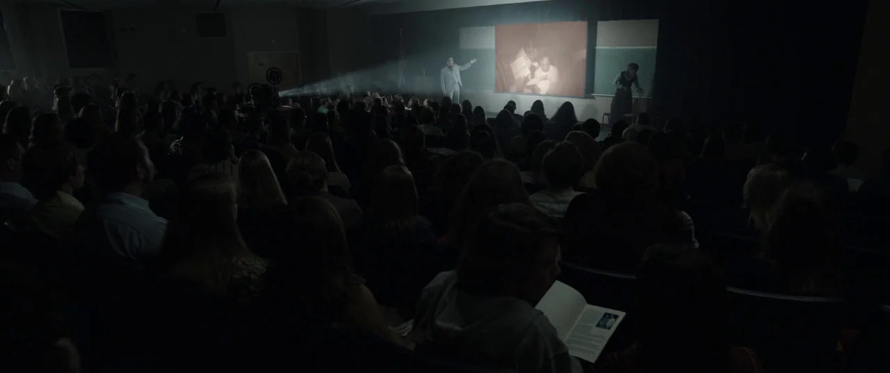 The Nun (2018) movie still: wide — A dark, tiered auditorium fills with an audience watching a presentation on a screen. A man stands on stage in…