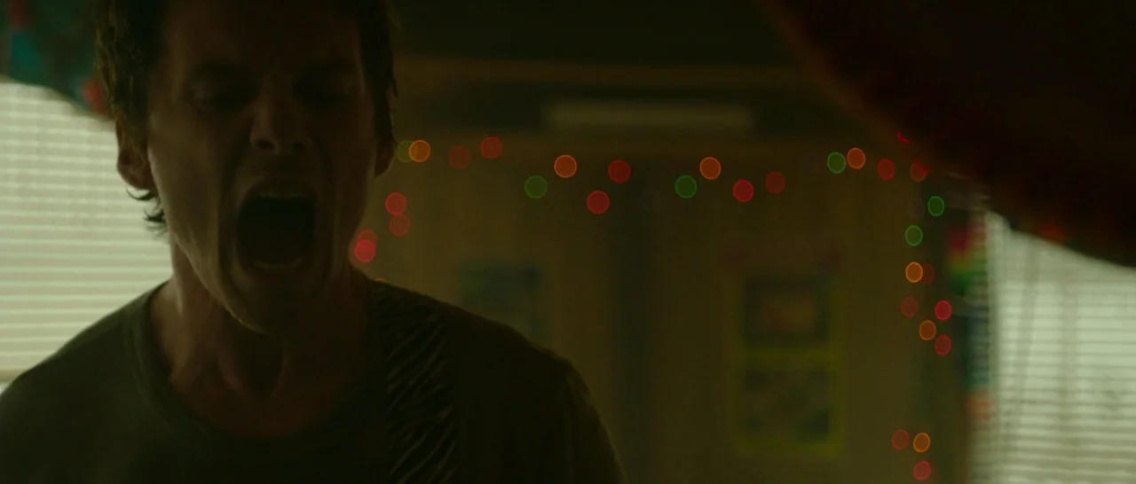 Green Room (2015) movie still: medium — An interior space is dimly lit by strings of multicolored lights creating bokeh in back. A man with brown…