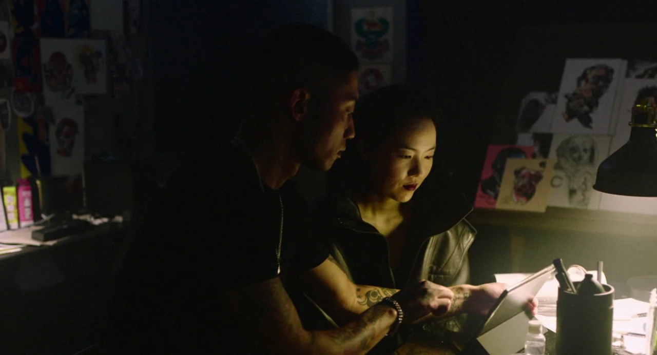 Return To Seoul (2022) movie still: medium — A dimly lit studio features walls covered in sketches and reference art drawings. A man and woman lean…