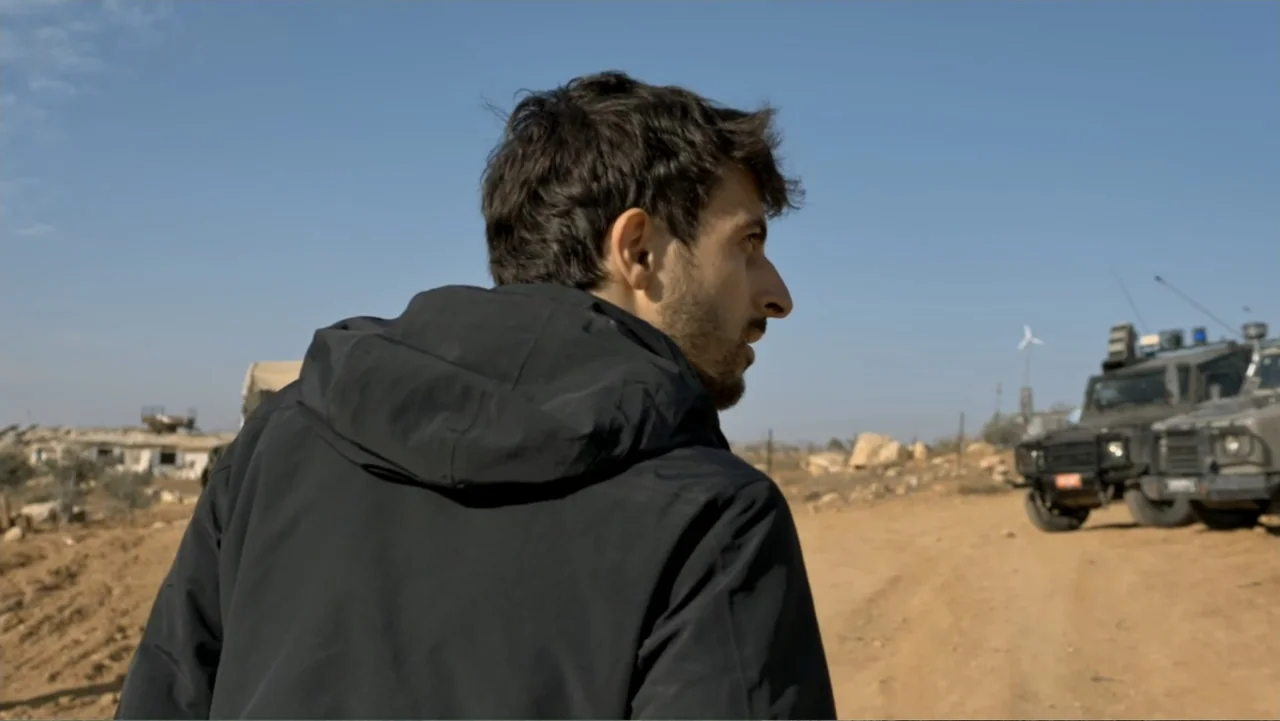 No Other Land (2024) movie still: medium — An arid dirt road in a desert landscape features military vehicles in the background. A man in a black…