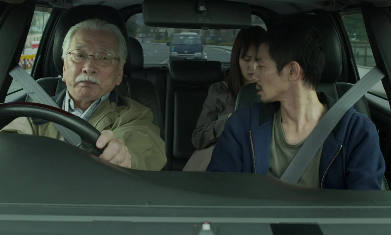 Like Someone In Love (2012) movie still: medium — Inside a moving vehicle, three people sit in the front and back seats. An elderly man drives while…
