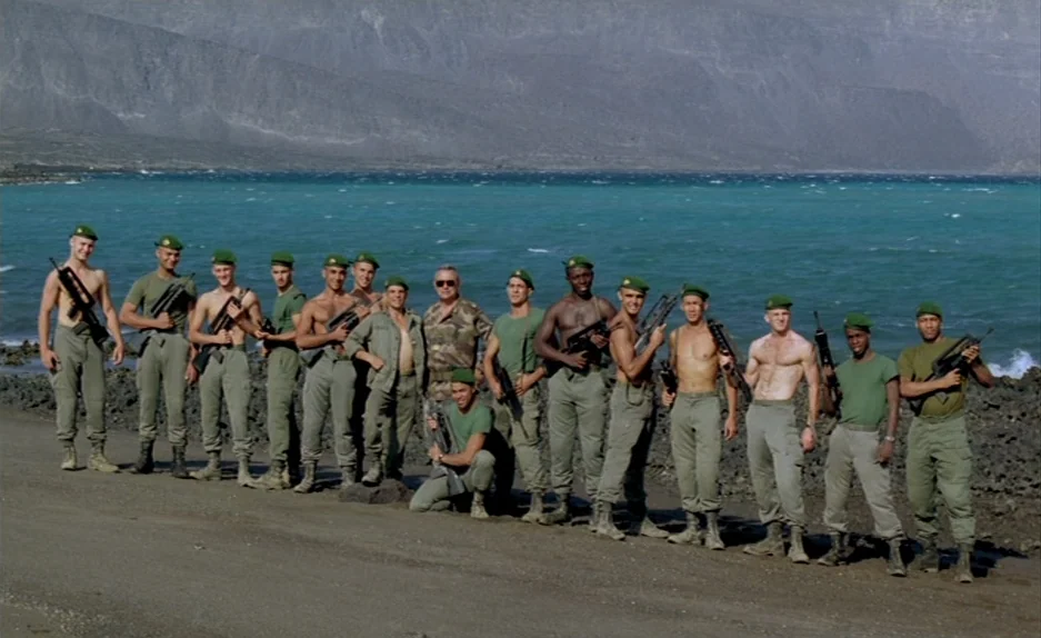 Beau Travail (1999) movie still: wide — A group of soldiers in green uniforms and berets stands on a coastal road. They are lined up against a…