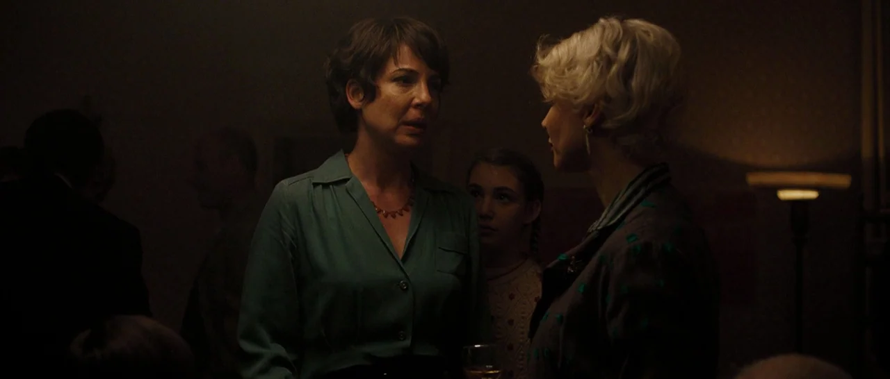 Pawn Sacrifice (2014) movie still: medium — A dimly lit room serves as the setting for a social gathering. Two women talk to each other while a…