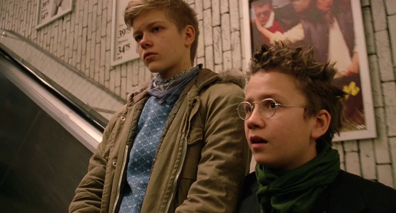 We Are The Best! (2013) movie still: medium — Two boys stand on a moving escalator in front of a white tiled wall. Advertising posters are mounted…