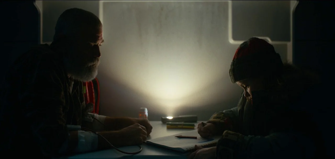 The Midnight Sky (2020) movie still: medium — A dimly lit, narrow metallic cabin features a small white table between two people. An elderly man and…