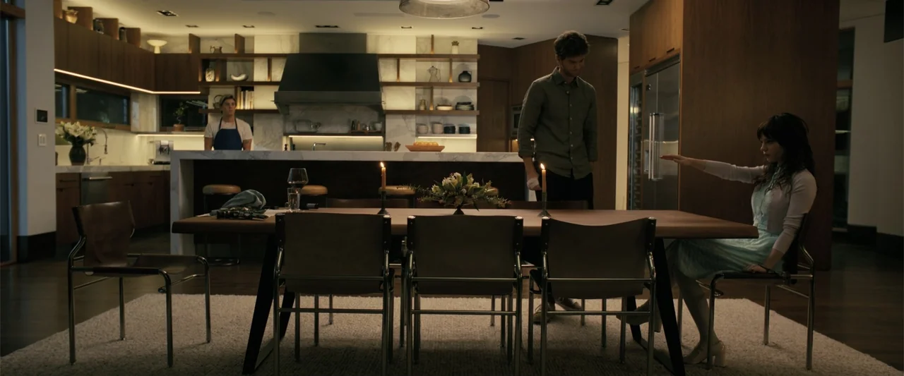 Companion (2025) movie still: wide — A spacious modern home interior features a large wooden dining table and open-concept cabinetry. A man stands…