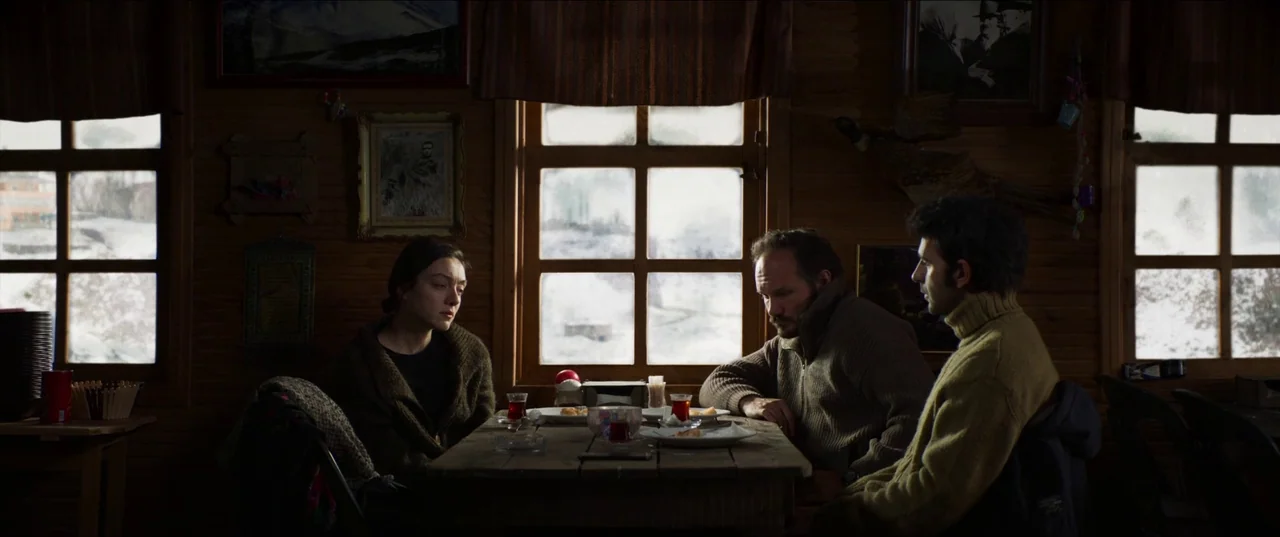 About Dry Grasses (2023) movie still: medium — A wooden rustic interior with framed photos and windows looking out onto snow. Three people sit at a…