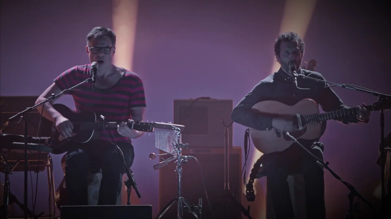 Flight of the Conchords: Live in London (2018) movie still: medium — A dimly lit indoor music stage featuring two men performing in front of…