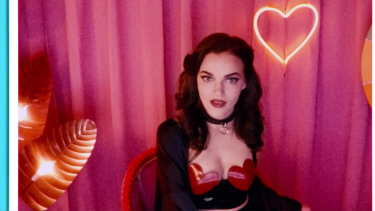 Cam (2018) movie still: medium — A small booth with pink curtains and hanging neon heart lights. A woman in a dark outfit sits facing the camera.