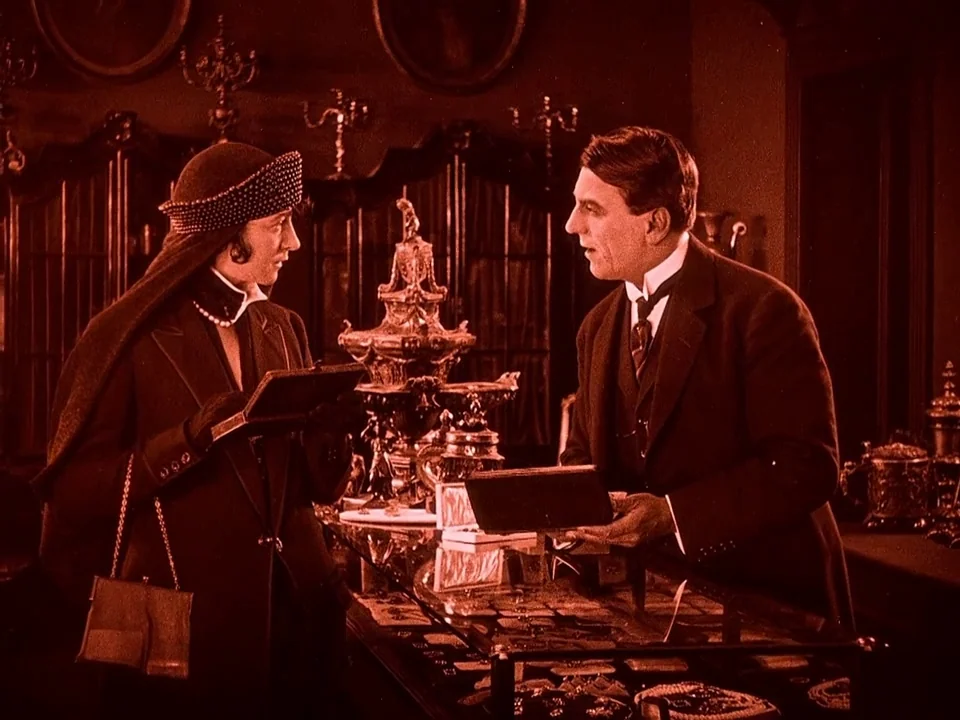 Häxan (1922) movie still: medium — A dimly lit 1920s store interior features ornate glass display cases filled with small items. A clerk stands…