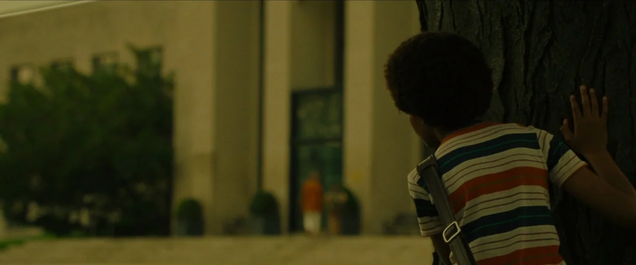 Wonderstruck (2017) movie still: medium — A young boy stands partially hidden behind the rough bark of a large tree. He watches a blurred building…