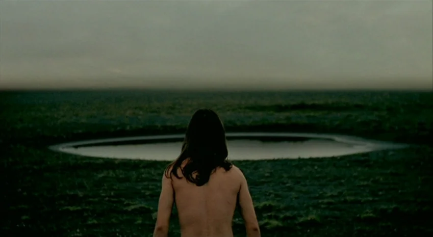 Rampo Noir (2005) movie still: medium — A flat, desolate field features a dark, circular pool of water in the distance. A woman with long hair…