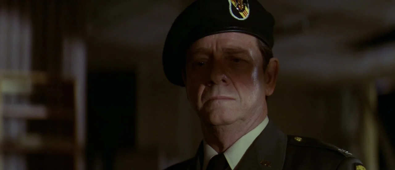 First Blood (1982) movie still: closeup — A dark military setting with indistinct background features suggestive of a desk or office. A man wearing…