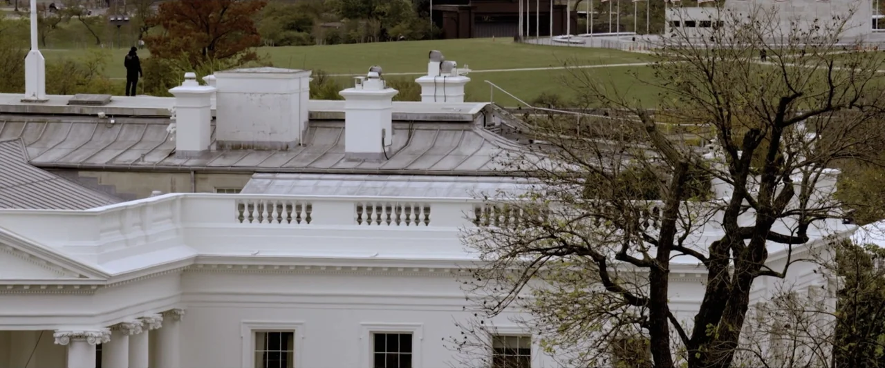 Reagan (2024) movie still: wide — The exterior roof and balustrade of a large white government building in late spring. A solitary individual in…