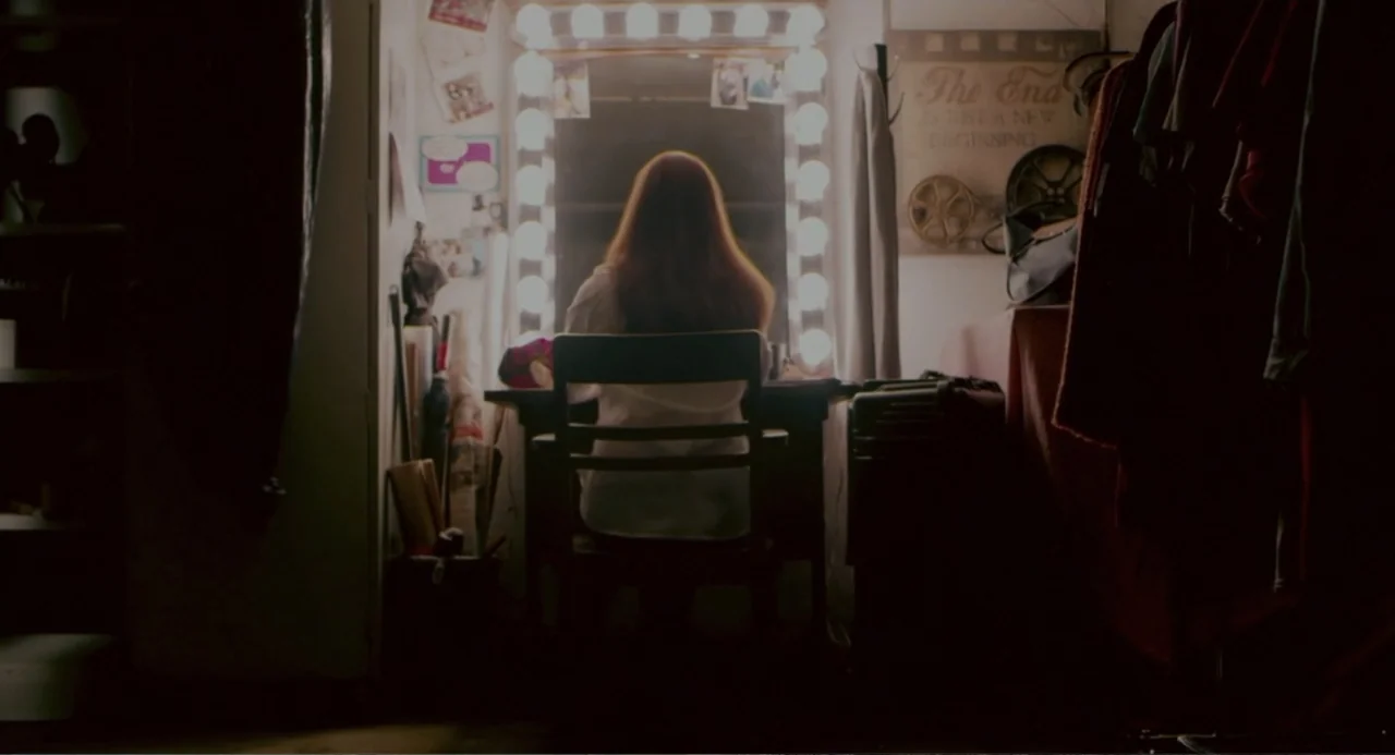 Terrifier (2018) movie still: medium — A cluttered dressing room vanity is illuminated by a ring of bright light bulbs. A person sits on a wooden…