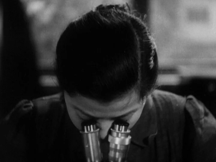 The Most Beautiful (1944) movie still: closeup — A dimly lit scientific space features a woman leaning over a bench. She gazes intently through the…