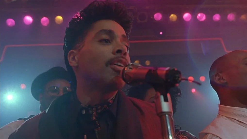 Graffiti Bridge (1990) movie still: medium — A dimly lit concert stage features vibrant pink and teal background lights. A male singer performs into…