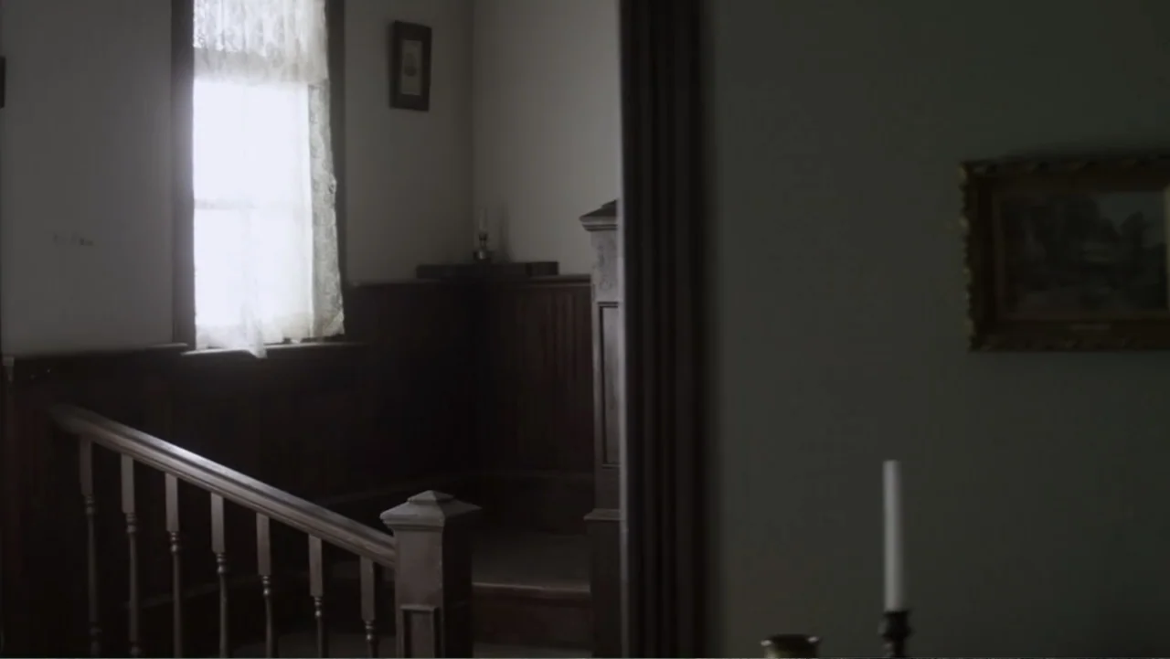 Dawn Rider (2012) movie still: wide — A dimly lit interior landing features wooden wainscoting and a staircase railing. A single window with lace…