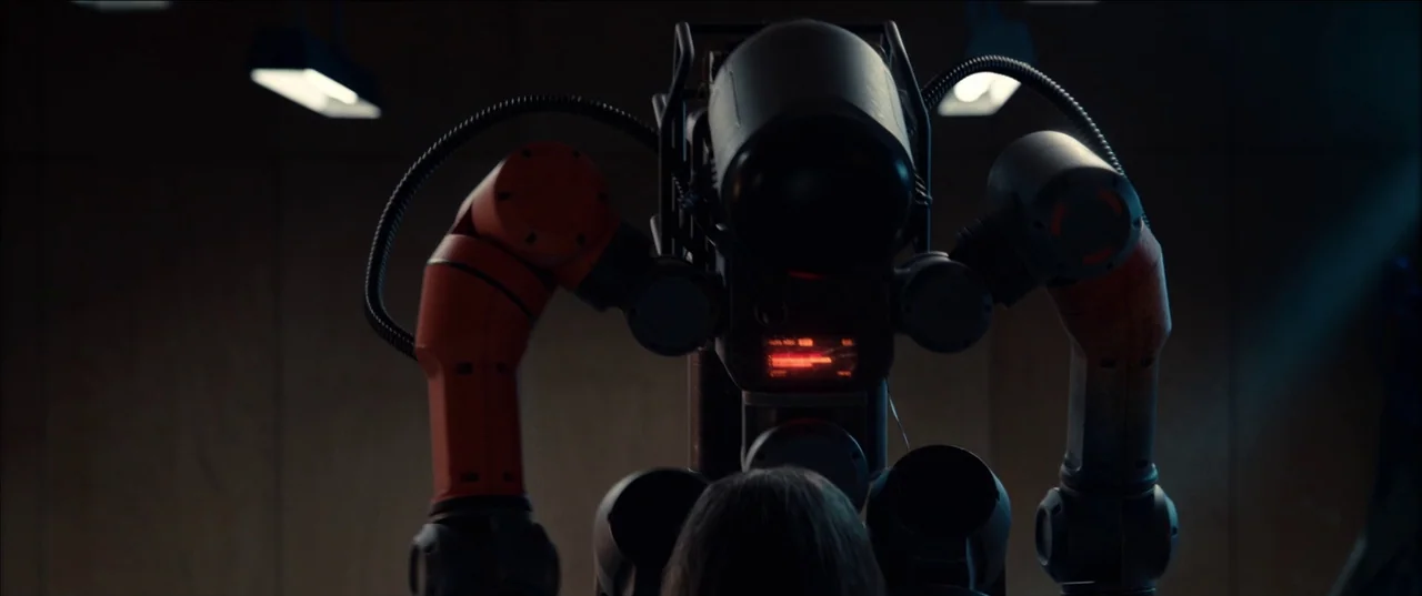 M3gan (2022) movie still: medium — A dimly lit room contains a large industrial robot with orange mechanical arms. A person stands with their back…