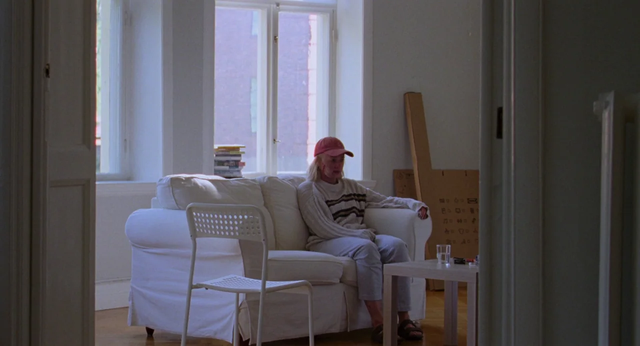 Sick of Myself (2022) movie still: medium — A living room interior contains a white sofa, a plastic chair, and a table. An elderly woman sits on the…