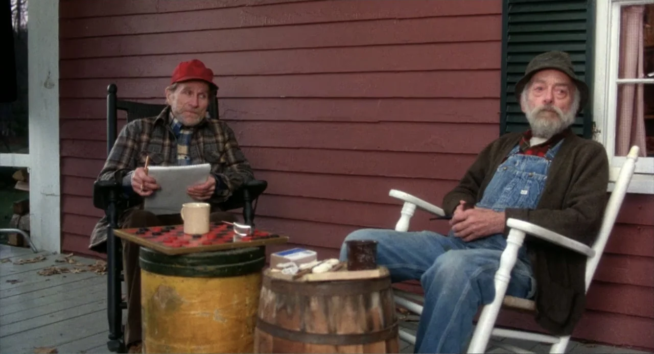 Baby Boom (1987) movie still: medium — A wooden porch with red siding features two rocking chairs and a barrel table. Two elderly men sit together…