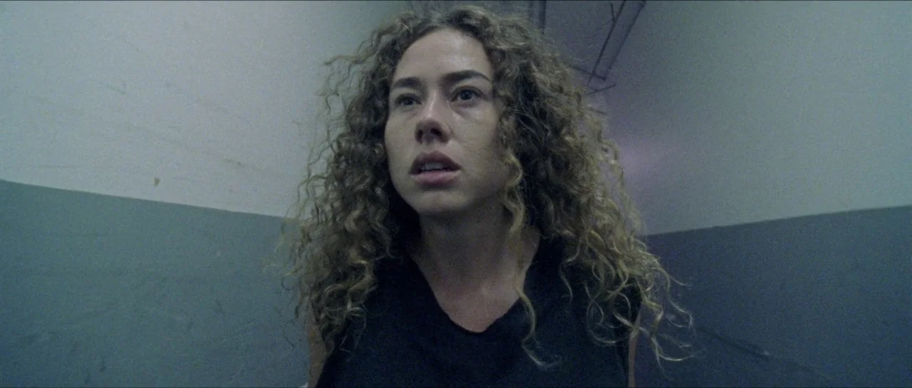 Bliss movie still: closeup — A dimly lit concrete stairwell with two-toned grey walls and exposed ceiling infrastructure. A woman with curly hair…