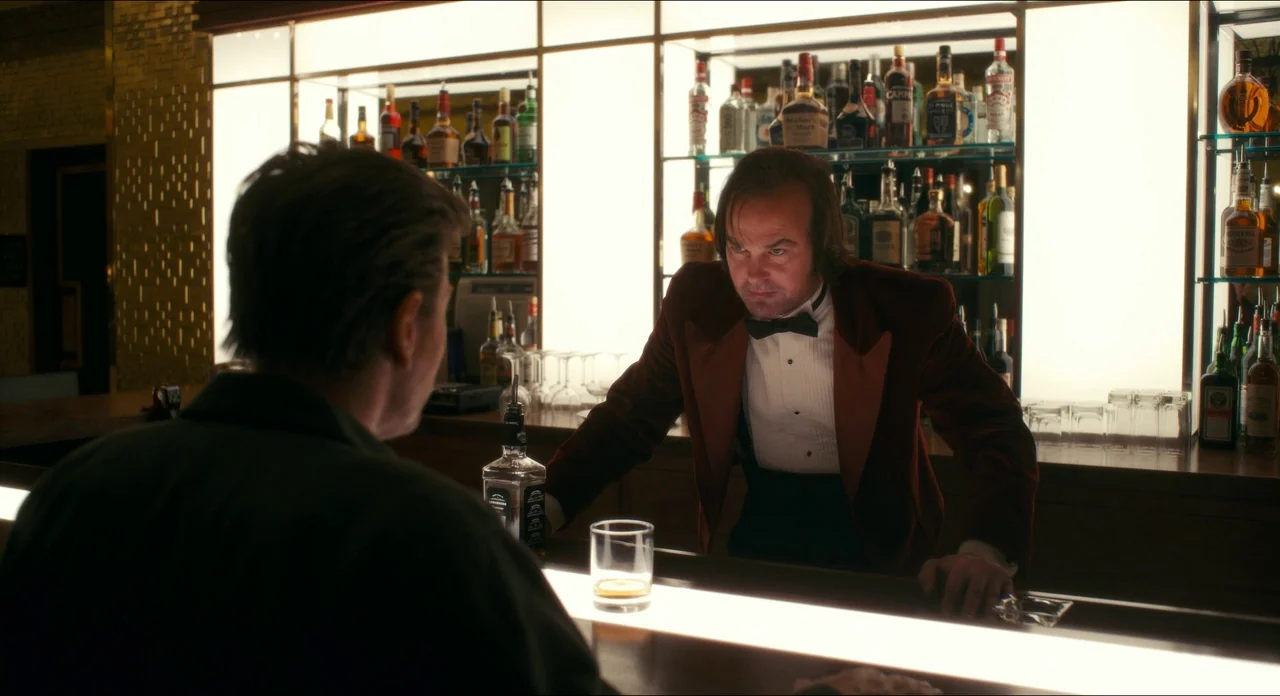 Doctor Sleep movie still: medium — A dimly lit room features a polished wooden bar with backlit glass shelving displays. A bartender in a red velvet…