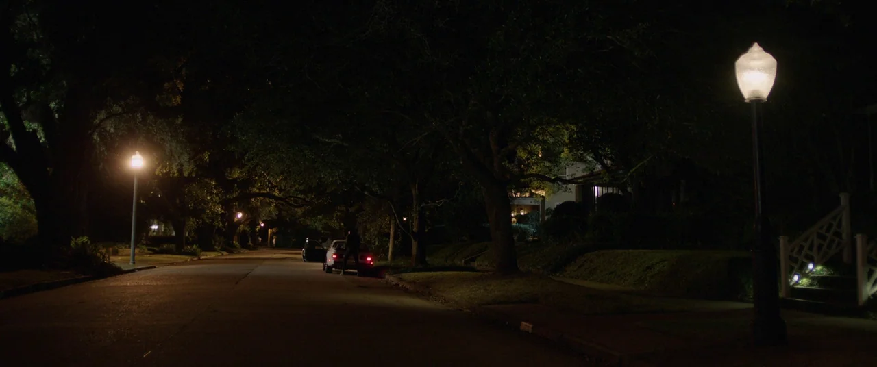 Get Out (2017) movie still: wide — A quiet residential street at night lined with large trees and old-fashioned lamps. A single dark vehicle sits…