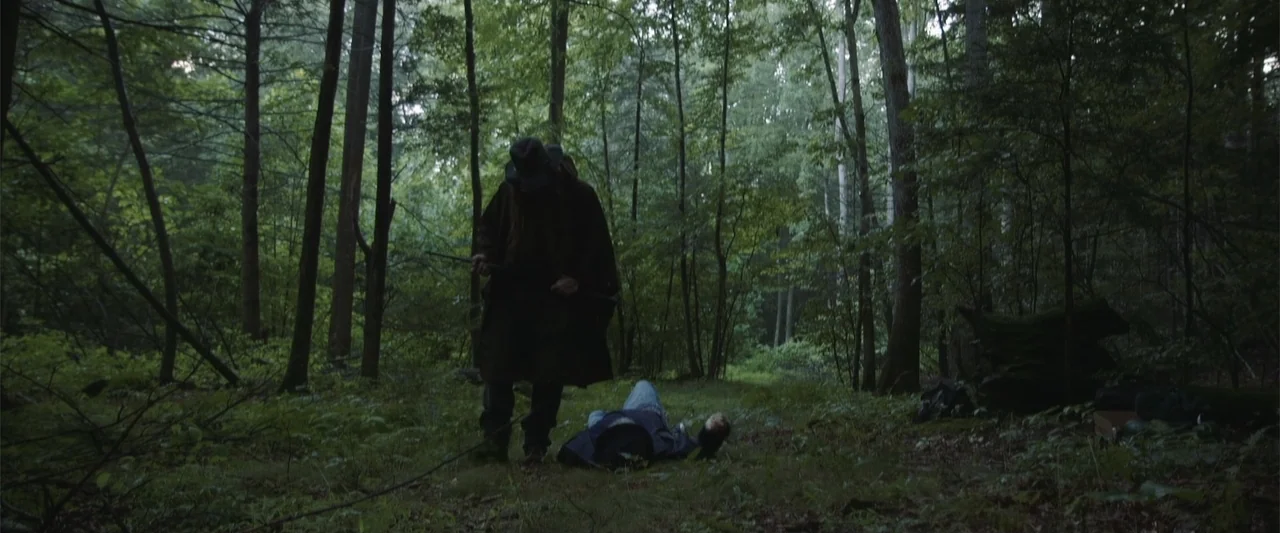 Darkness Waits (2020) movie still: wide — A dense patch of temperate woodland features tall trees and low ground vegetation. One man stands over a…