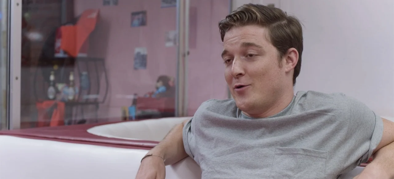 Hellbreeder (2004) movie still: medium — A corner booth in a diner with pink walls and white padded seating. A young man wearing a grey t-shirt sits…