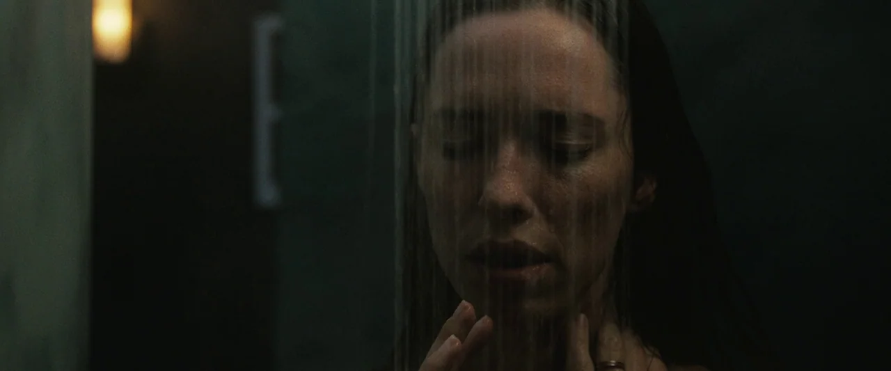 The Night House (2020) movie still: closeup — A dimly lit shower stall features dark tiles and water falling in vertical streams. A woman stands…