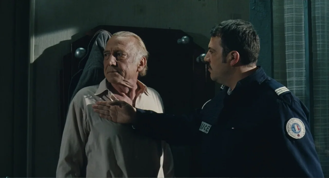 Le Havre (2011) movie still: medium — An interior space with a dark wooden wall unit and a window with curtain. A uniformed police officer holds his…