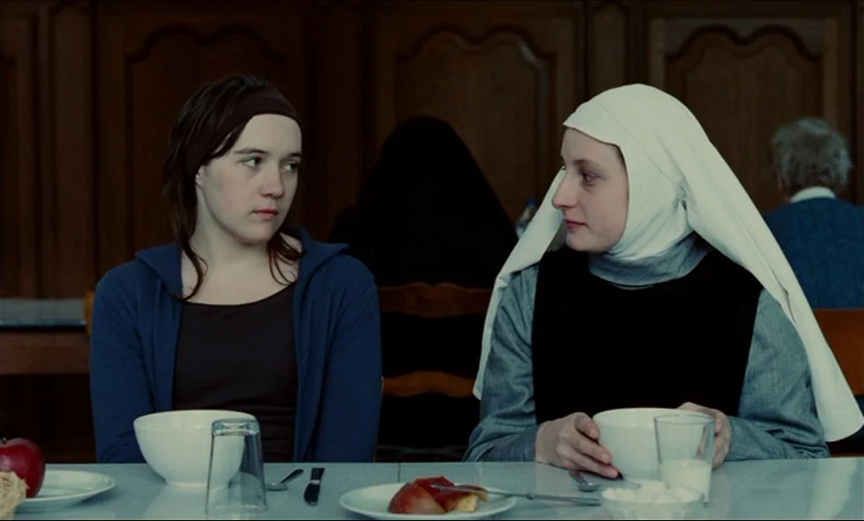 Hadewijch (2009) movie still: medium — Two women sit at a white table in a plain, wood-paneled room. The woman on the left wears casual clothing,…
