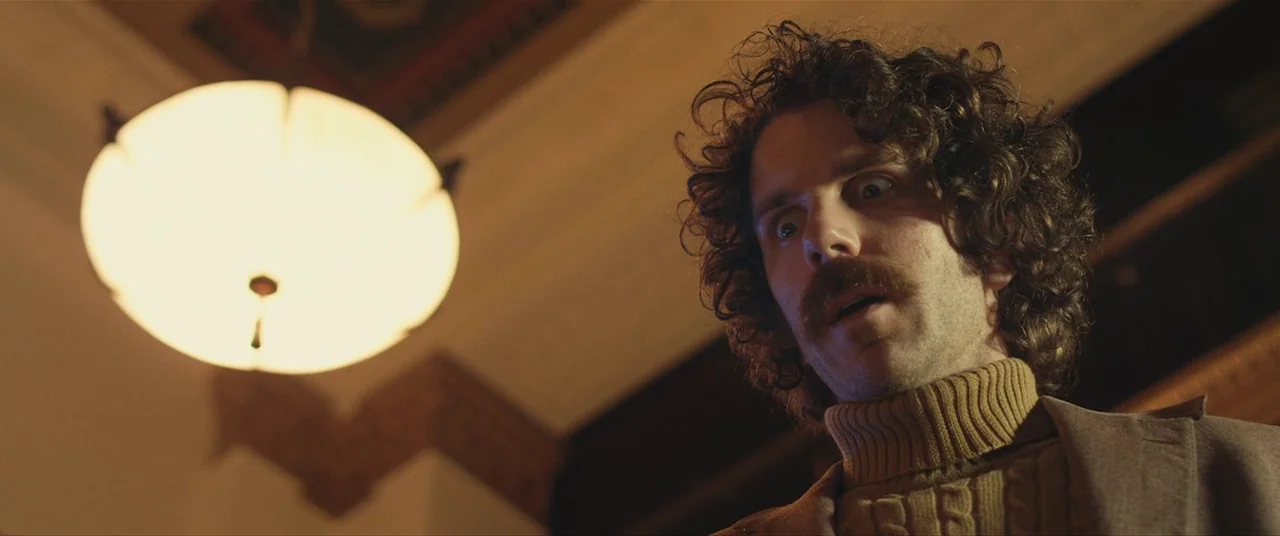 The Editor (2014) movie still: A man with a distinctive mustache and curly hair reacts to an unseen sight with wide-eyed shock, illuminated from…