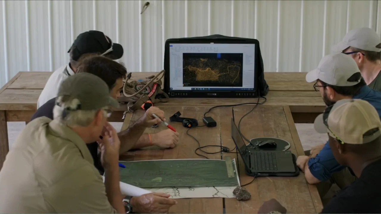 Finding Amelia (2024) movie still: medium — A group of people are gathered around a wooden table in a rustic room. They are looking at a computer…
