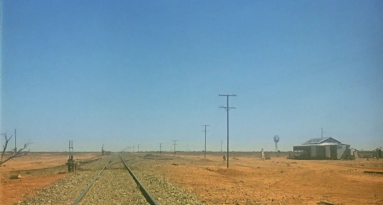 Wake In Fright (1971) movie still: establishing — A flat, arid desert landscape features a set of train tracks receding into distance. A small,…