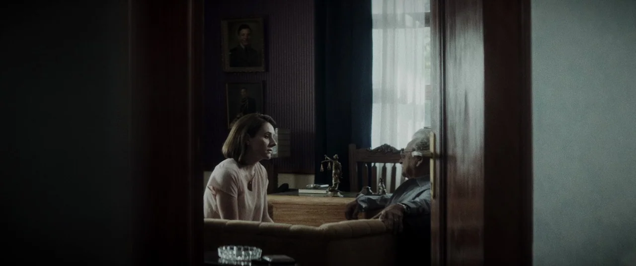 La Llorona (2019) movie still: medium — A dark study with deep purple walls decorated with framed portraits of men. A middle-aged woman sits facing…