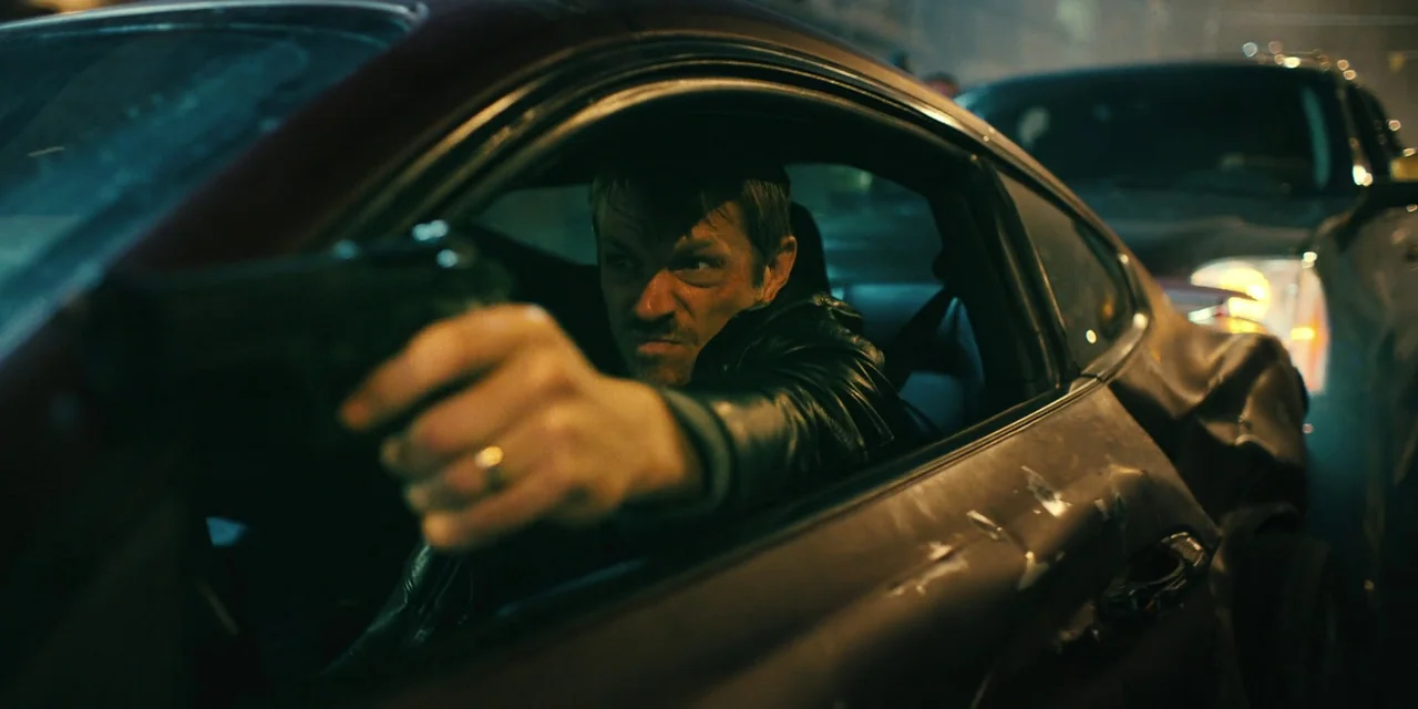 Silent Night (2023) movie still: medium — A man sits inside a dark vehicle at night while holding a handgun outward. The scene is illuminated by…